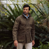 Man wearing a brown Tom Beckbe Tensaw Jacket and light pants stands outdoors with green plants behind him.