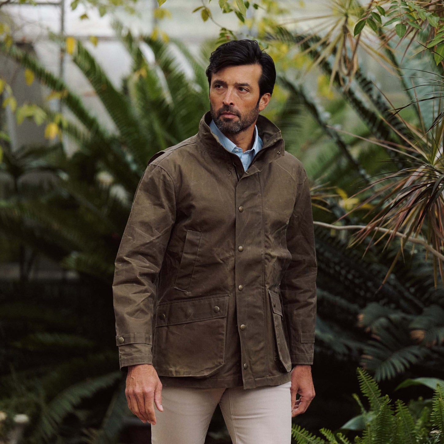 Man wearing a brown Tom Beckbe Tensaw Jacket and light pants stands outdoors with green plants behind him.