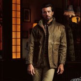 Man wearing a brown Tom Beckbe Tensaw Jacket and khaki pants stands indoors by a window in warm, ambient lighting.