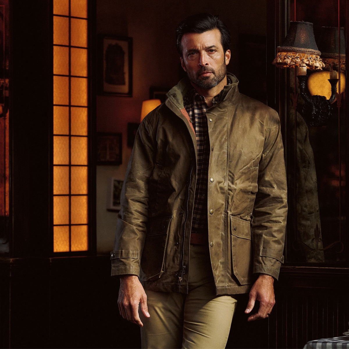 Man wearing a brown Tom Beckbe Tensaw Jacket and khaki pants stands indoors by a window in warm, ambient lighting.