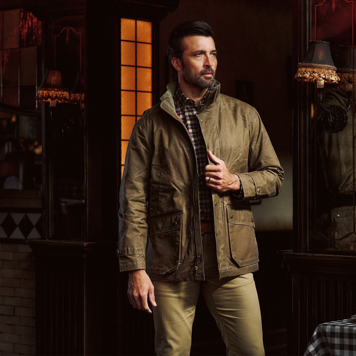 Man wears a brown Tom Beckbe Tensaw Jacket and khaki pants, standing indoors with one hand on his durable jacket.