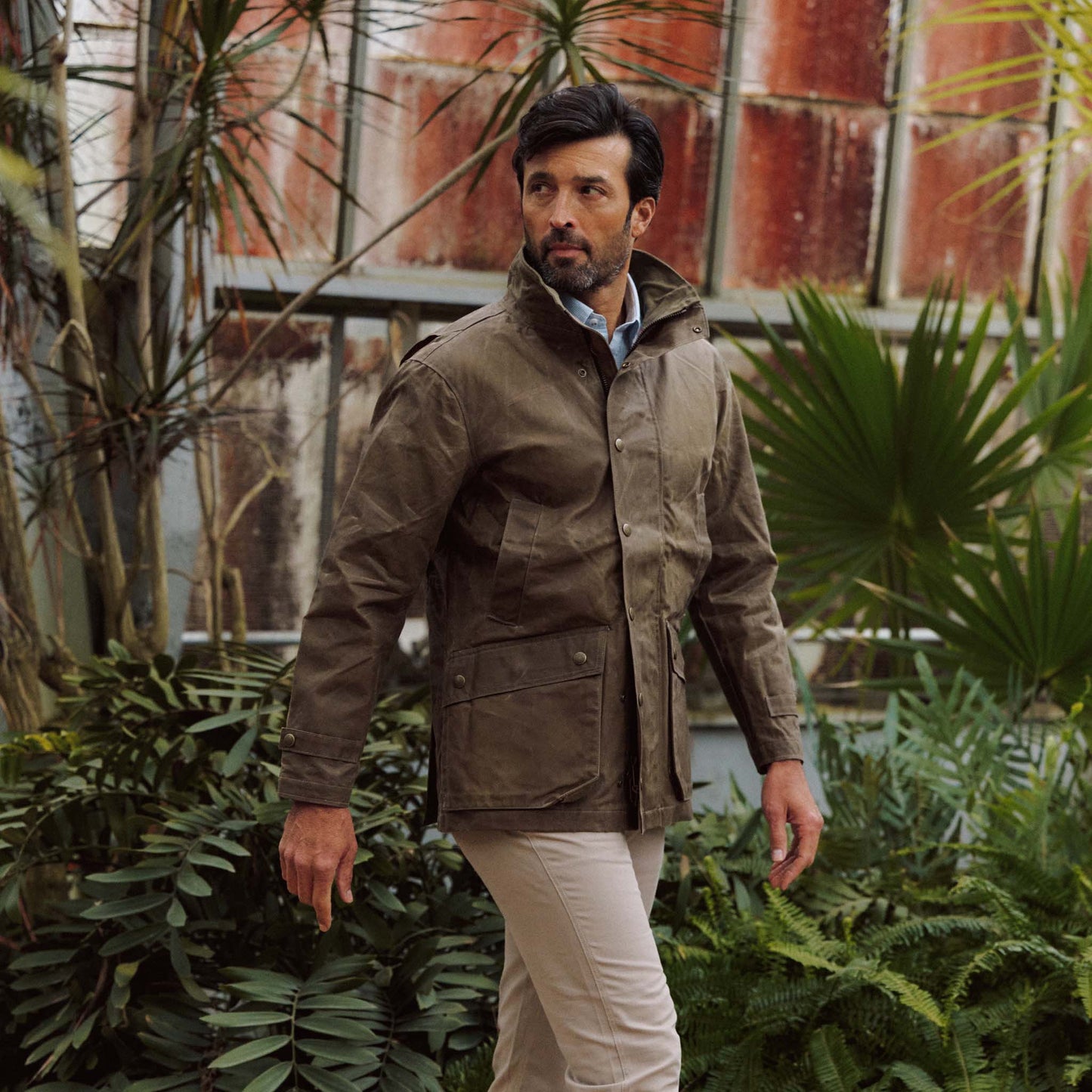 Man in a rugged Tom Beckbe Tensaw Jacket and beige pants stands amid green plants before a weathered greenhouse.