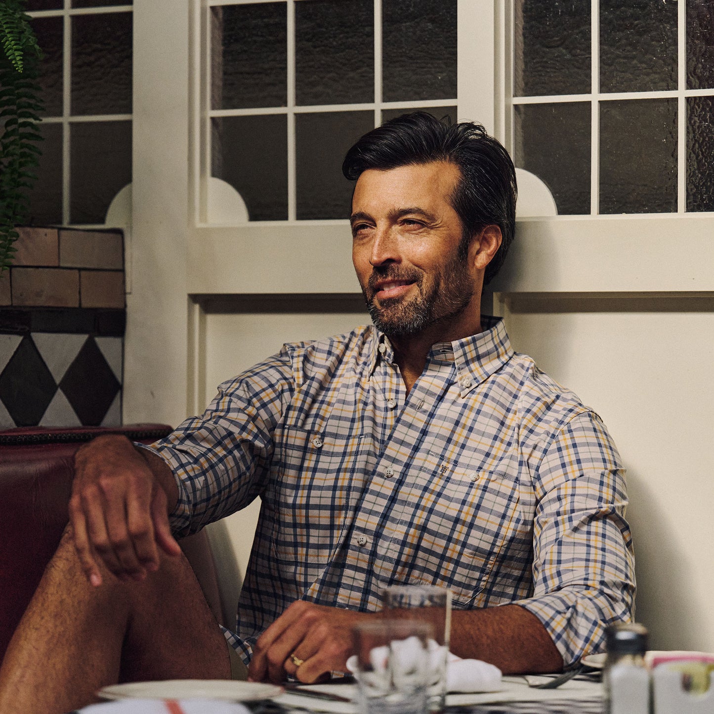 A man in a Tom Beckbe Tidewater Shirt smiles at a restaurant table with glassware and plates in front of him.