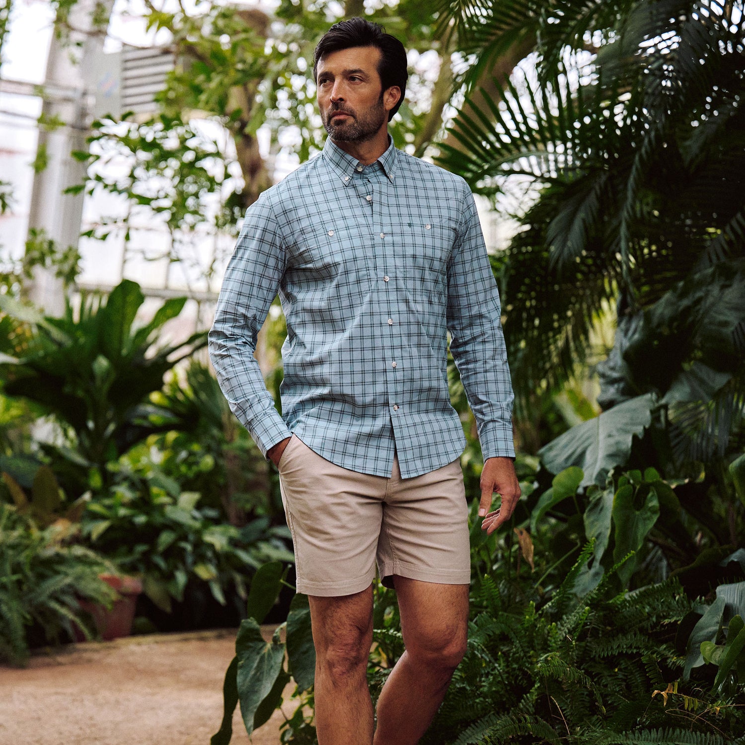Man wears Tom Beckbe Tidewater Shirt (blue plaid, UPF 30+) and beige shorts among green plants in a greenhouse.