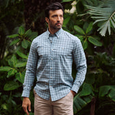 Man wears Tom Beckbe Tidewater Shirt and beige shorts, standing outdoors before lush green foliage.