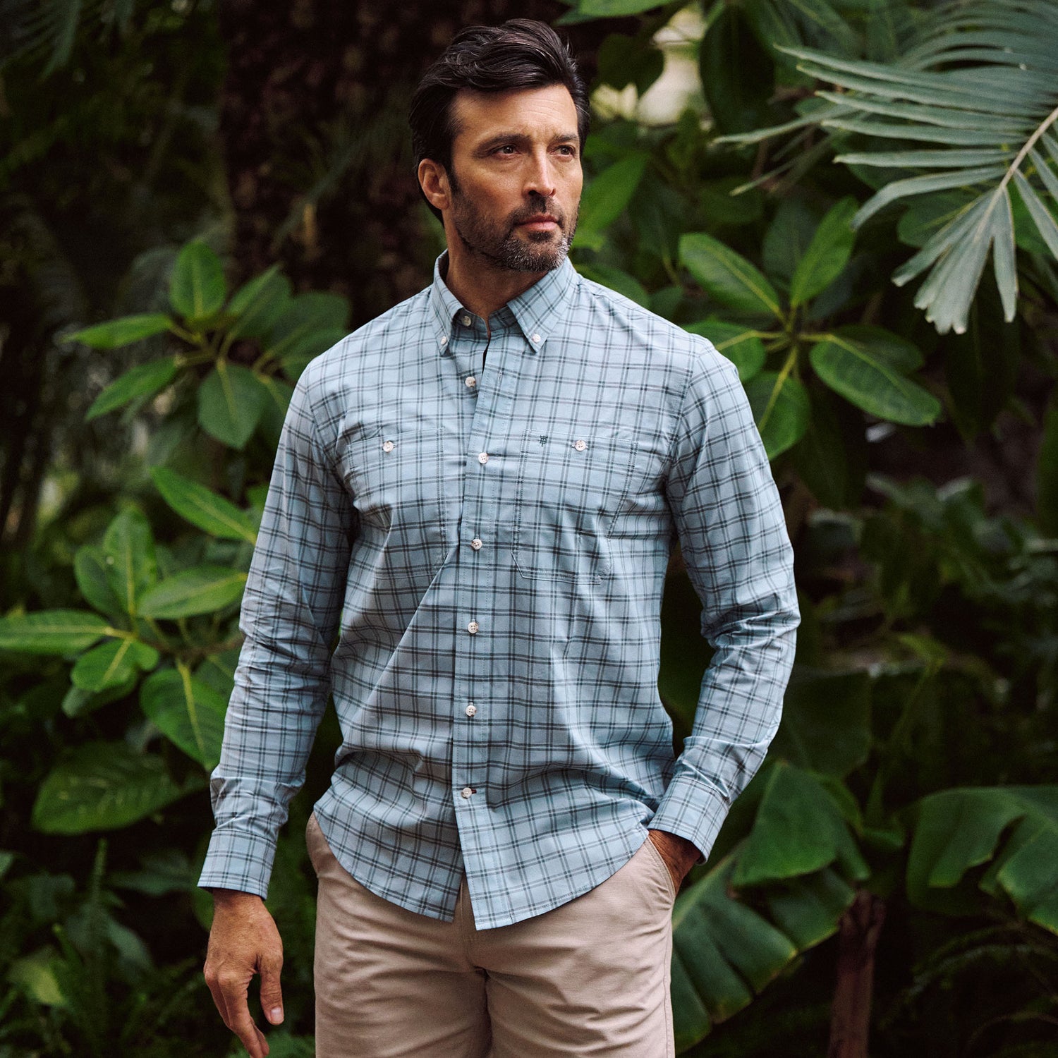 Man wears Tom Beckbe Tidewater Shirt and beige shorts, standing outdoors before lush green foliage.