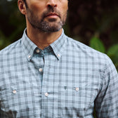 A man outdoors wears the Tom Beckbe Tidewater Shirt, a light blue plaid long sleeve, shown shoulders to mid-chest.
