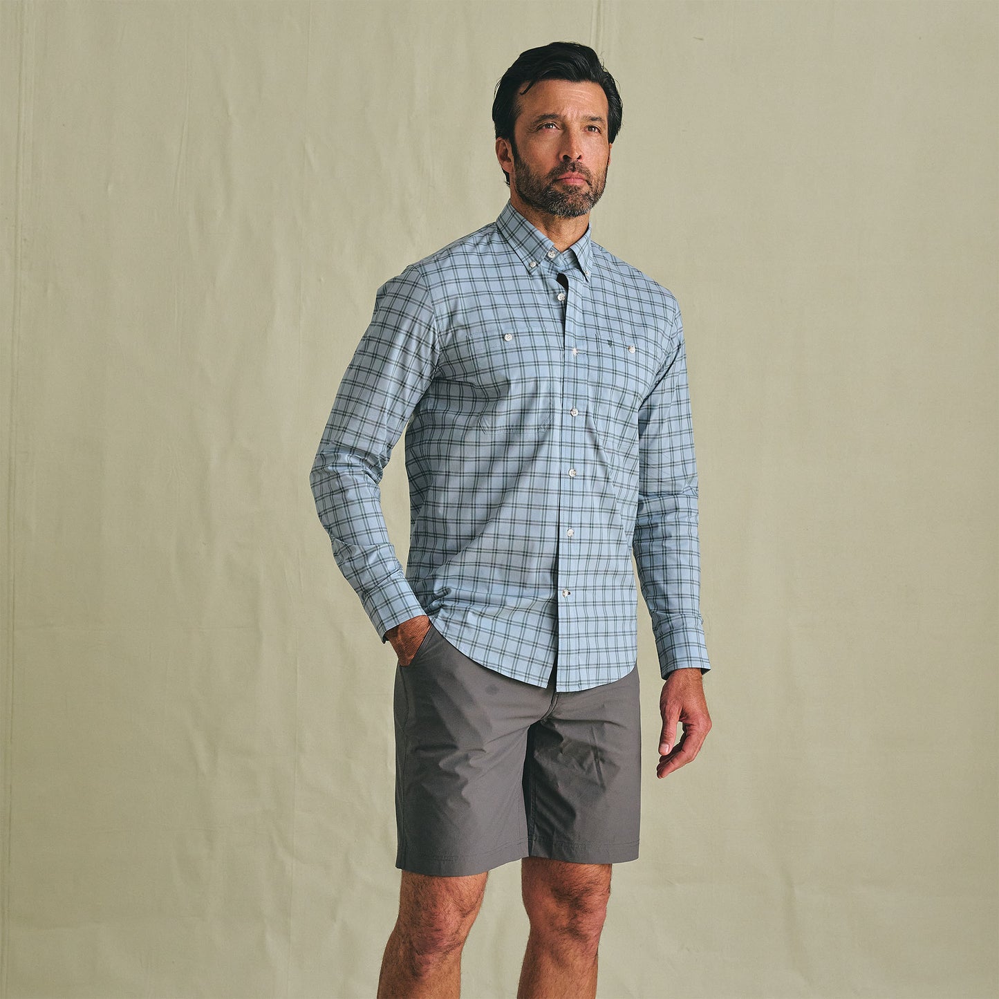 A man stands against a plain background wearing the Tom Beckbe Tidewater Shirt and gray shorts.