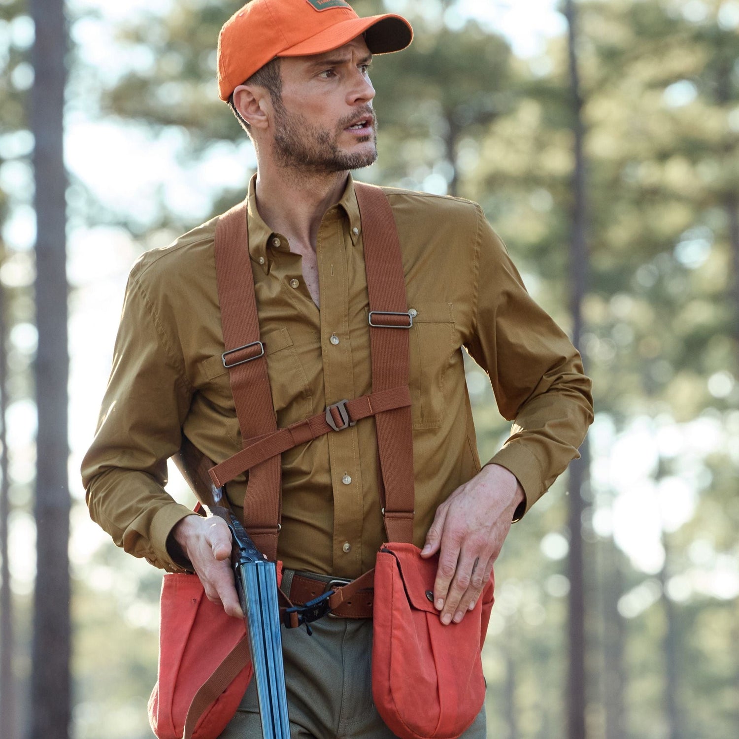 Man in a Tom Beckbe brown Tidewater Shirt with UPF 30+ holds a double-barreled shotgun, trees in the background.
