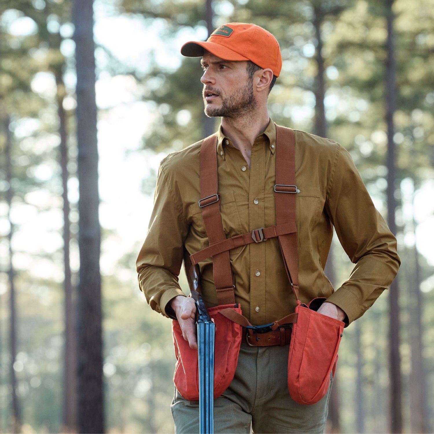 A man in an orange cap and Tidewater Shirt by Tom Beckbe walks through a forest with a rifle and UPF 30+ harness.