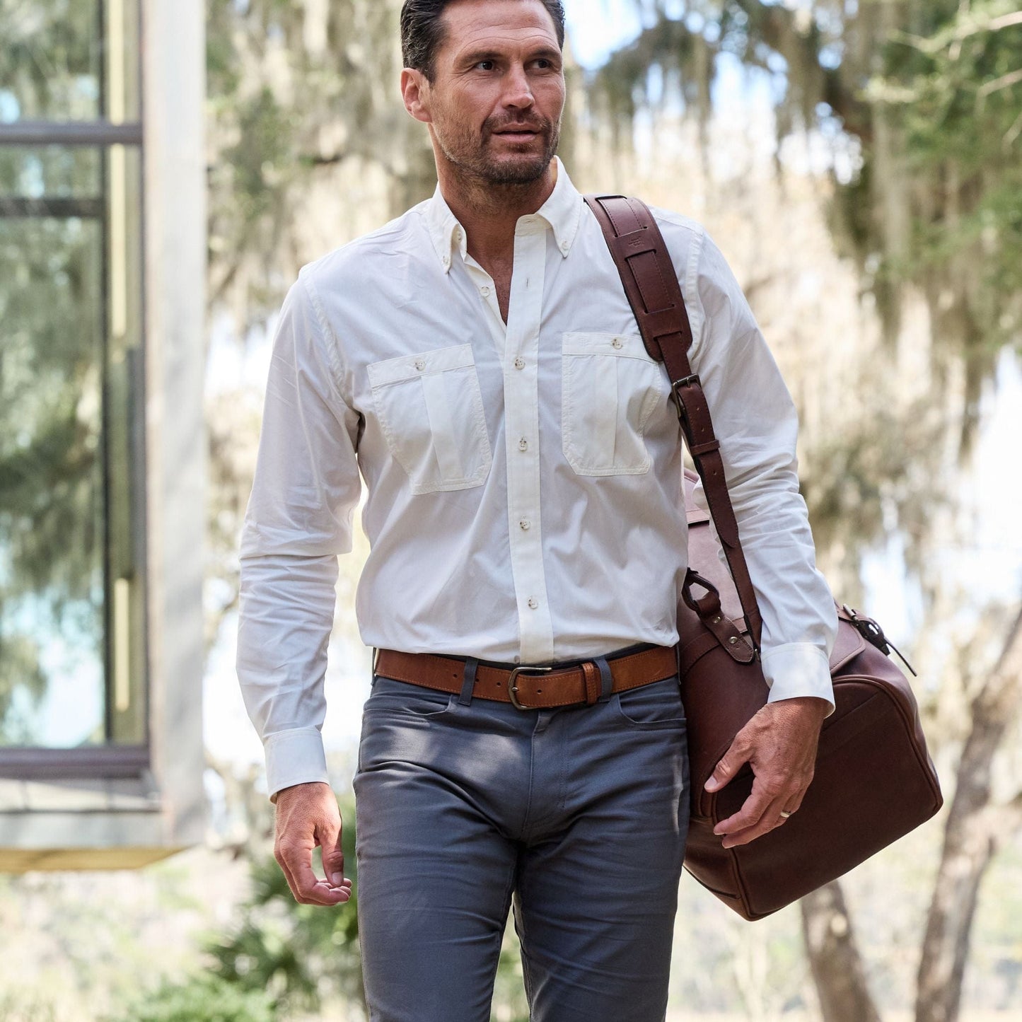 A man in a Tom Beckbe Tidewater Shirt walks outdoors, leather bag in hand, with trees and a window behind him.