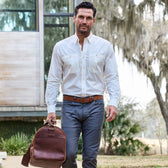 A man in a Tom Beckbe Tidewater Shirt and gray pants strolls outside with a brown bag under tree shade.