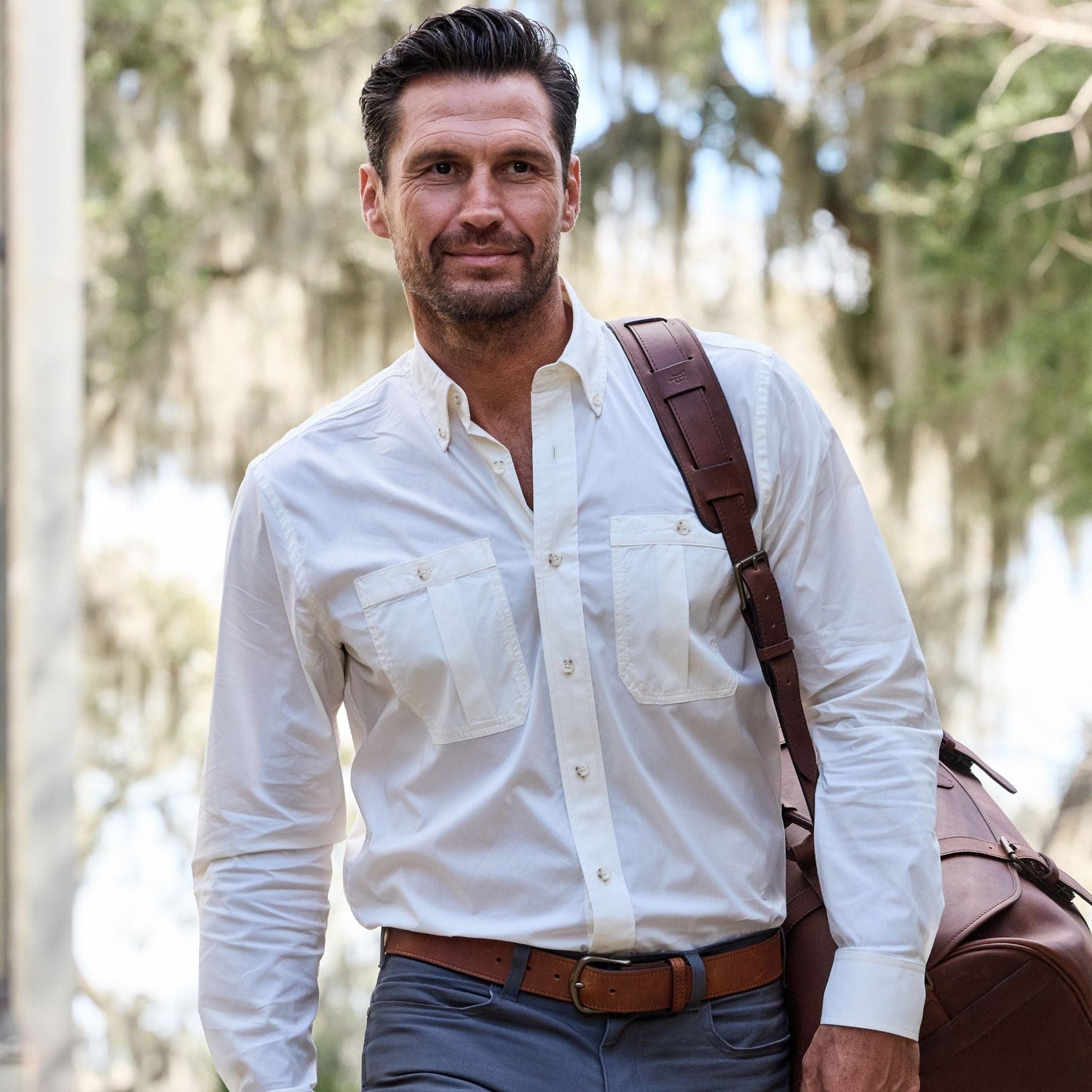 A man in a Tidewater Shirt by Tom Beckbe carries a large brown leather bag over his shoulder outdoors.