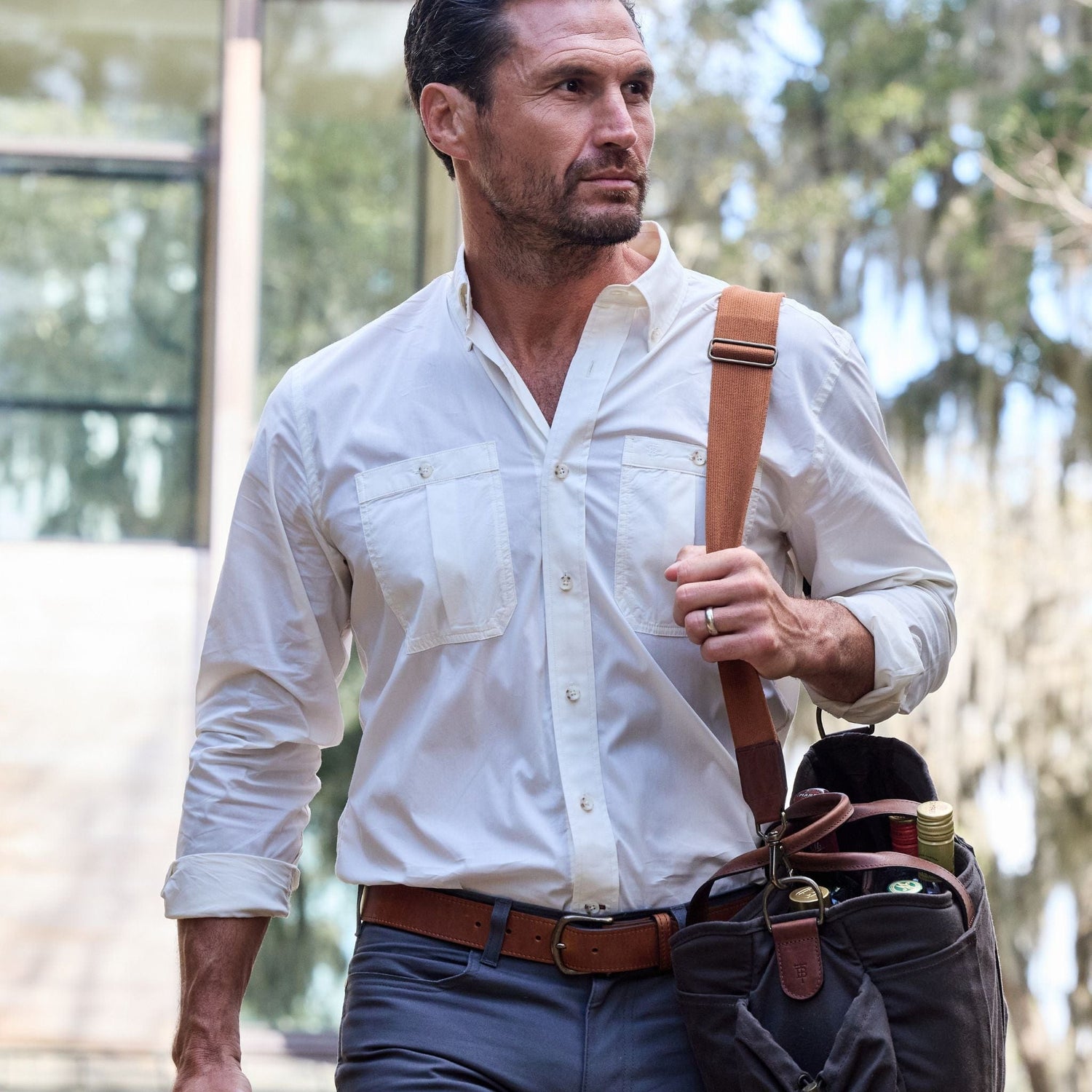 A man in a Tom Beckbe Tidewater Shirt carries a brown bag with bottles, standing near a building with large windows.