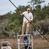 Man in a hat atop a boats tower, wearing a Tom Beckbe Tidewater Shirt, with marshy vegetation and trees behind.