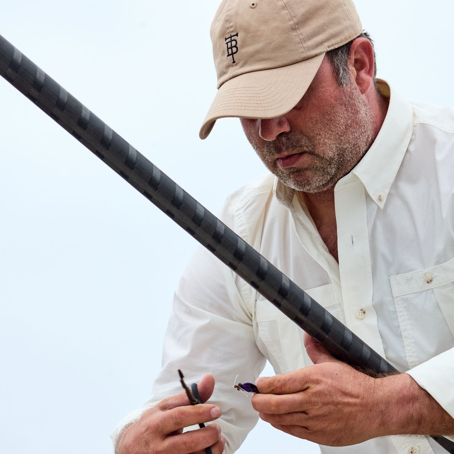 A man in a Tom Beckbe Tidewater Shirt and beige cap focuses on attaching a lure to his fishing rod.