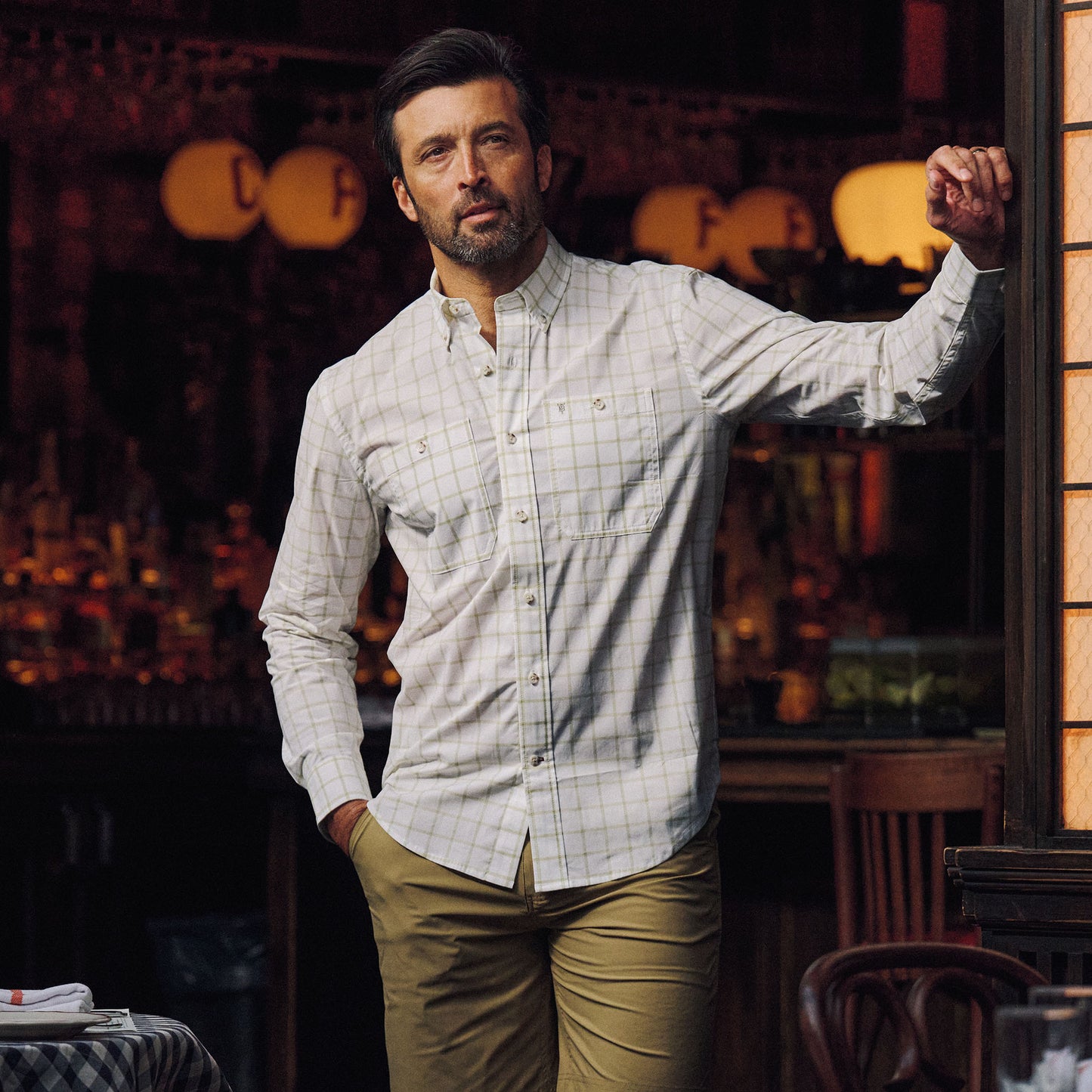 A man wears the Tom Beckbe Tidewater Shirt and khaki pants, leaning on a wooden frame in dim indoor light.