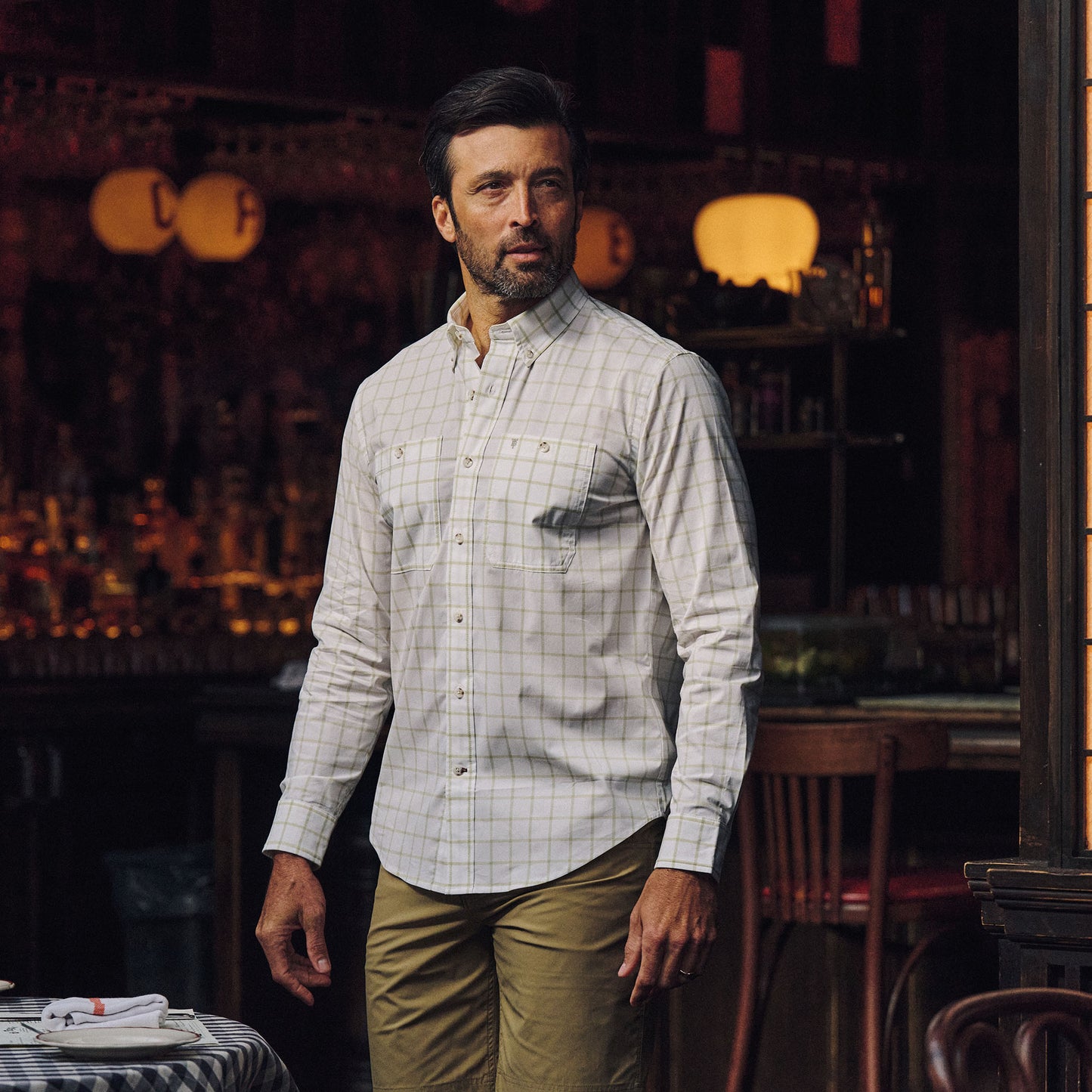 A man in a Tom Beckbe Tidewater Shirt and tan pants stands indoors near a table, with a dimly lit bar behind him.