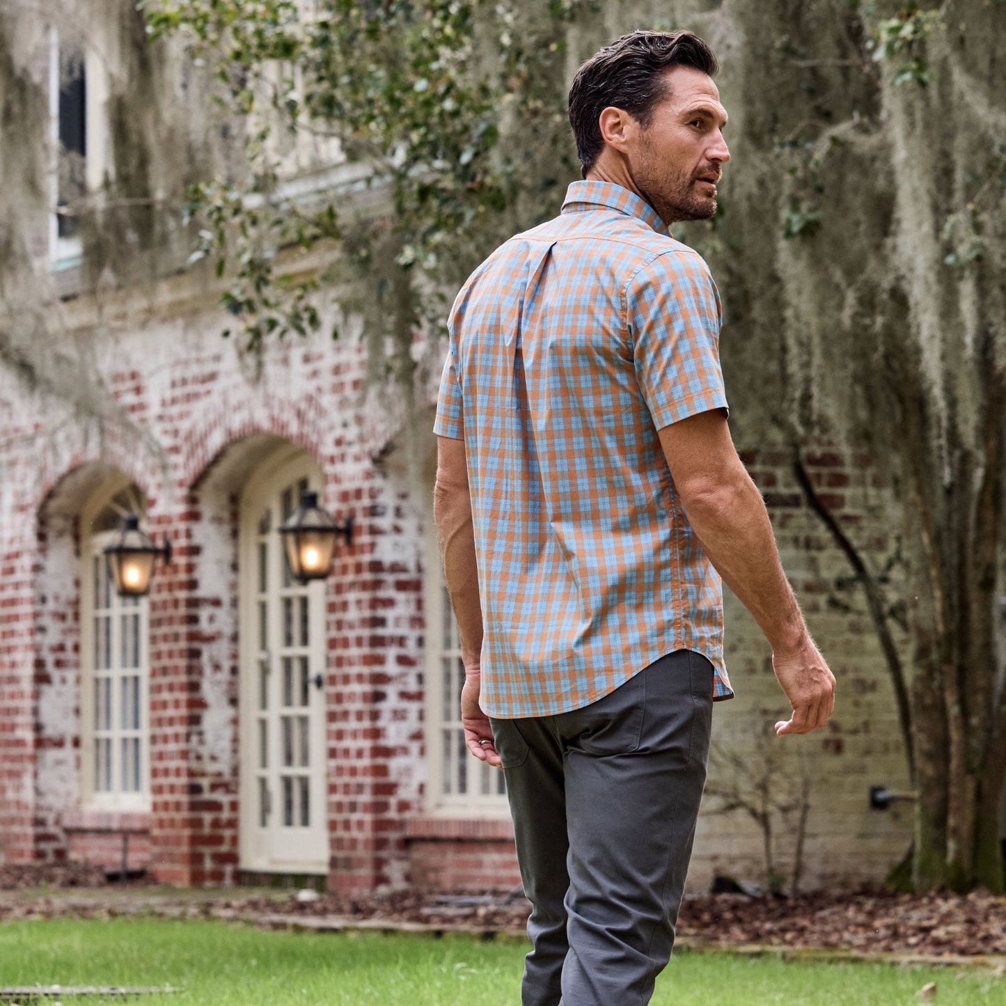 A man in a Tom Beckbe Tidewater Shirt strolls outside a brick building with arched windows and mossy trees.