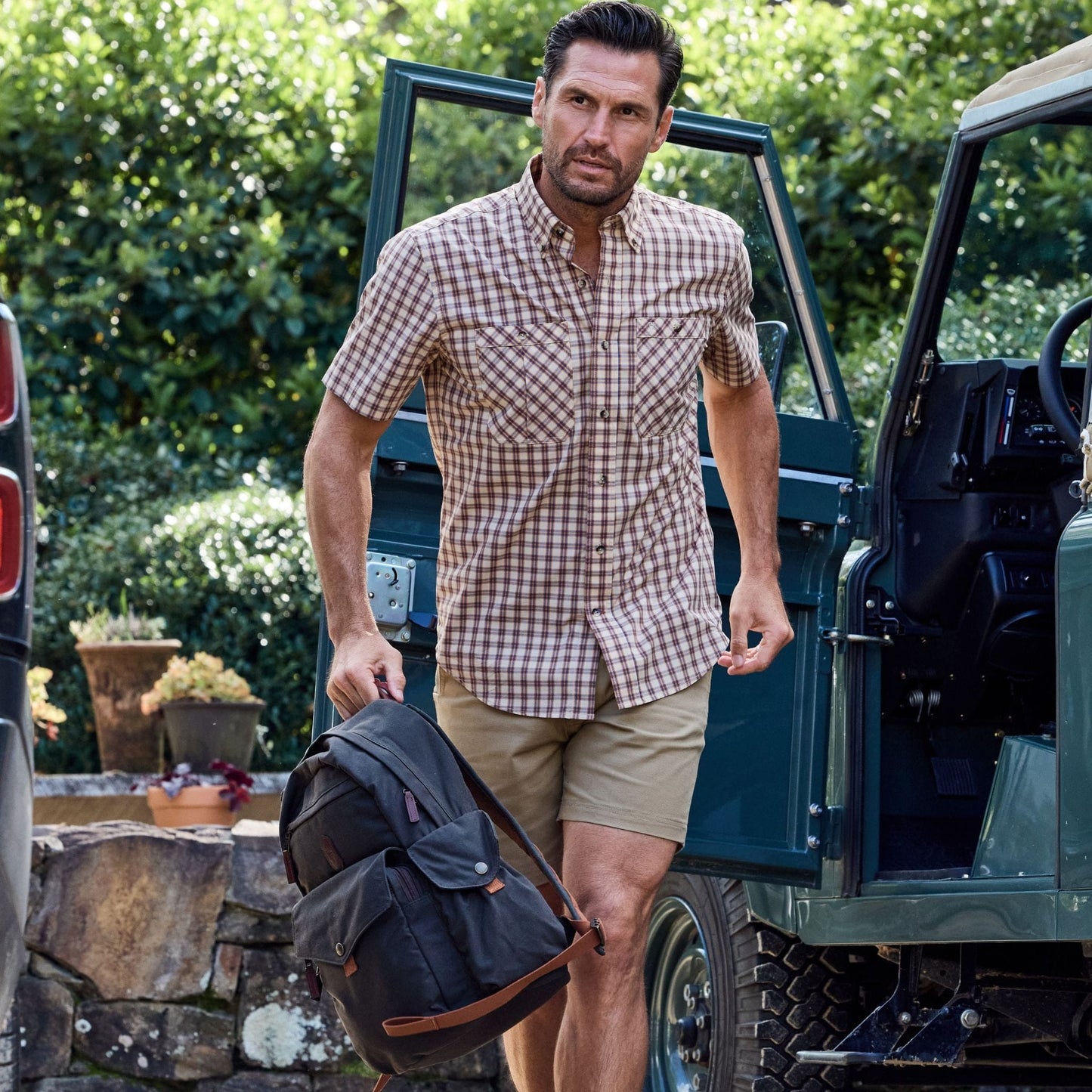 A man in a Tom Beckbe Tidewater Shirt stands by an open car door, wearing shorts, surrounded by lush greenery.