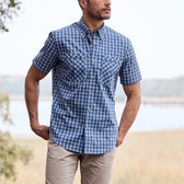 A man is outdoors in a Tom Beckbe Tidewater Shirt and beige pants, near water and trees.