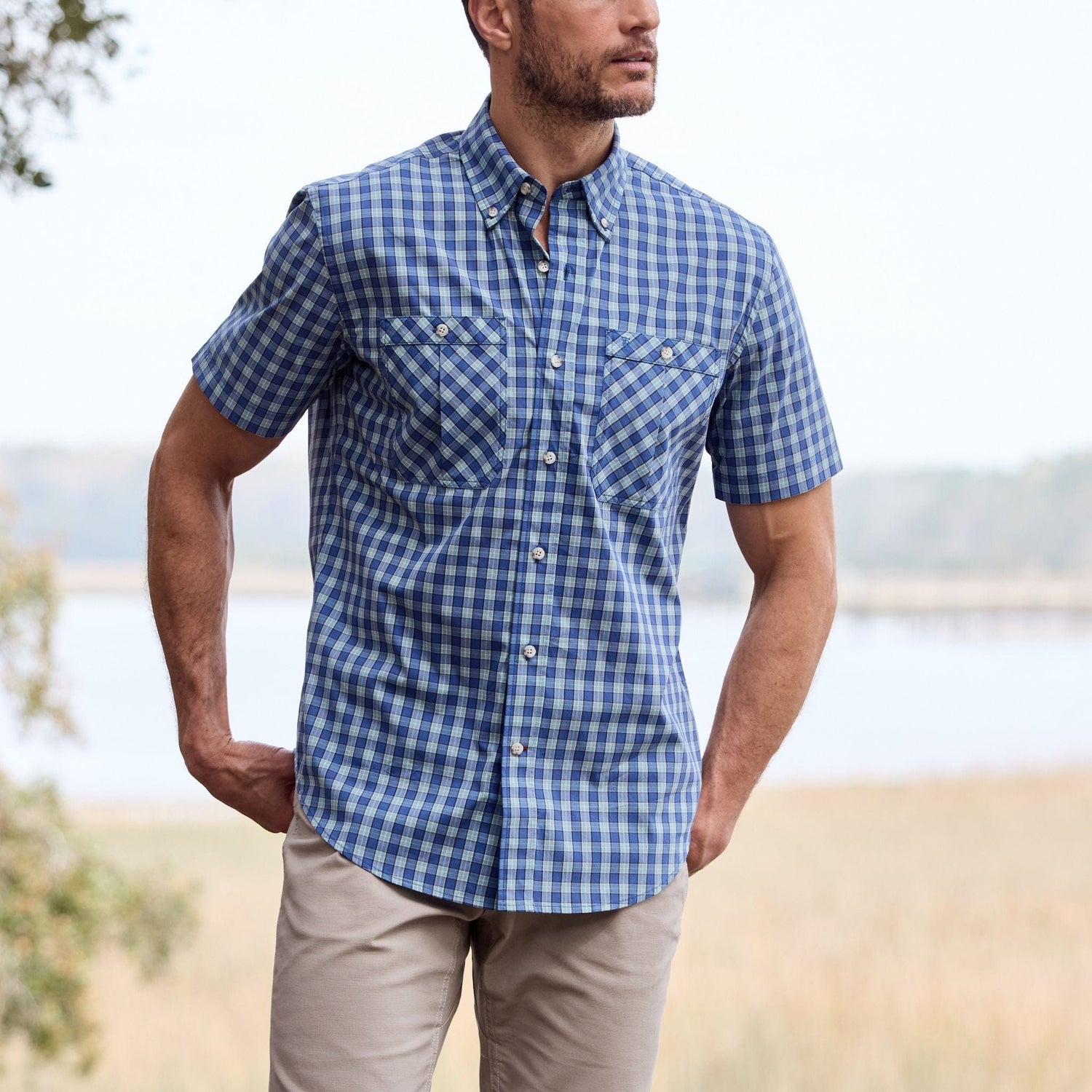 A man is outdoors in a Tom Beckbe Tidewater Shirt and beige pants, near water and trees.