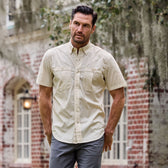 Man in a beige Tom Beckbe Tidewater Shirt and gray pants stands in front of a brick building with arched windows.