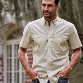 A man outside a brick building with hands in pockets wears a beige, short-sleeve Tidewater Shirt by Tom Beckbe with UPF 30+.