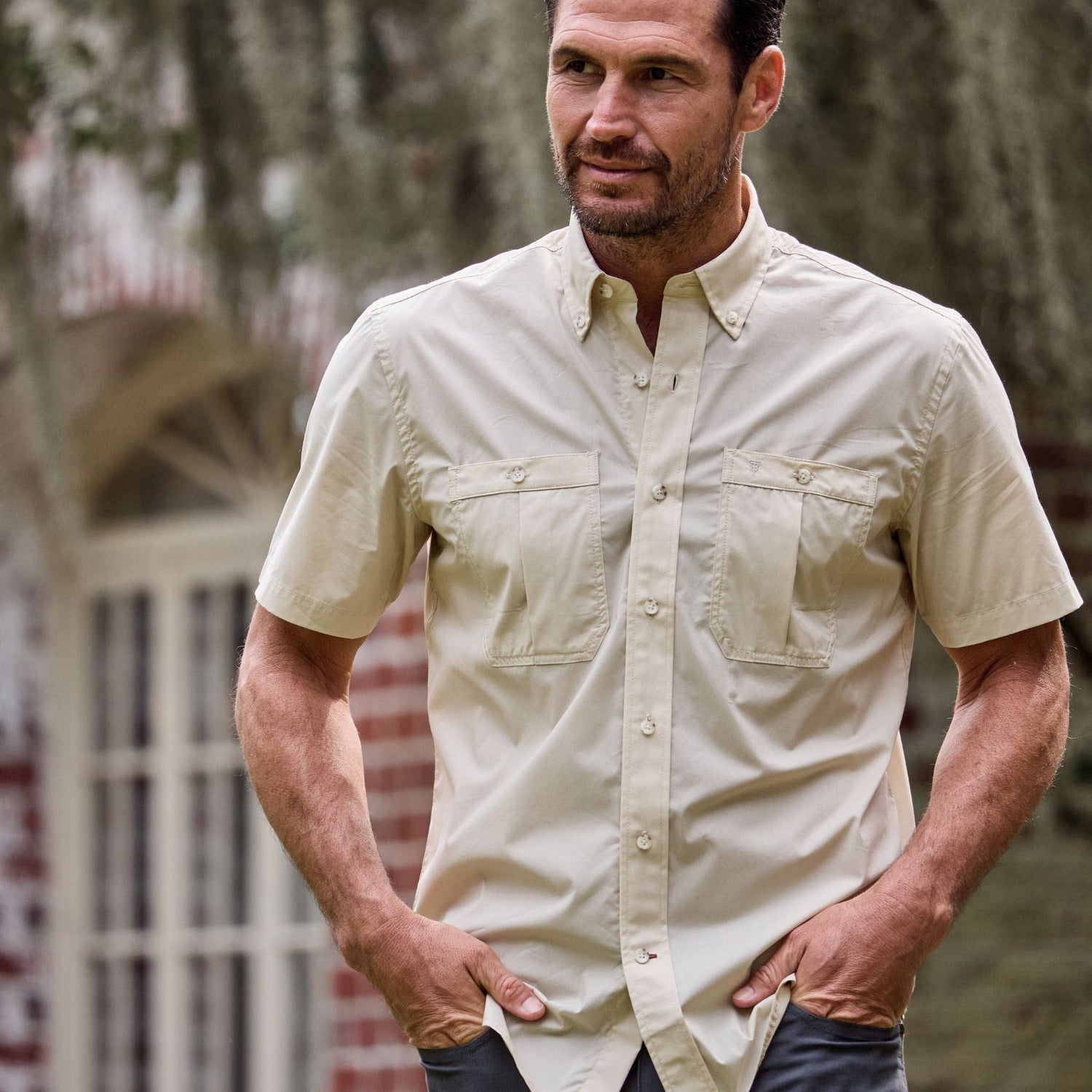 A man outside a brick building with hands in pockets wears a beige, short-sleeve Tidewater Shirt by Tom Beckbe with UPF 30+.