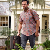 A man in a Tom Beckbe Tidewater Shirt carries a suitcase and shoulder bag, with a house in the background.