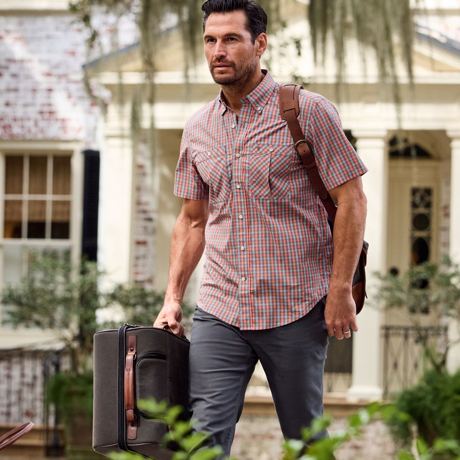 A man in a Tom Beckbe Tidewater Shirt carries a suitcase and shoulder bag, with a house in the background.