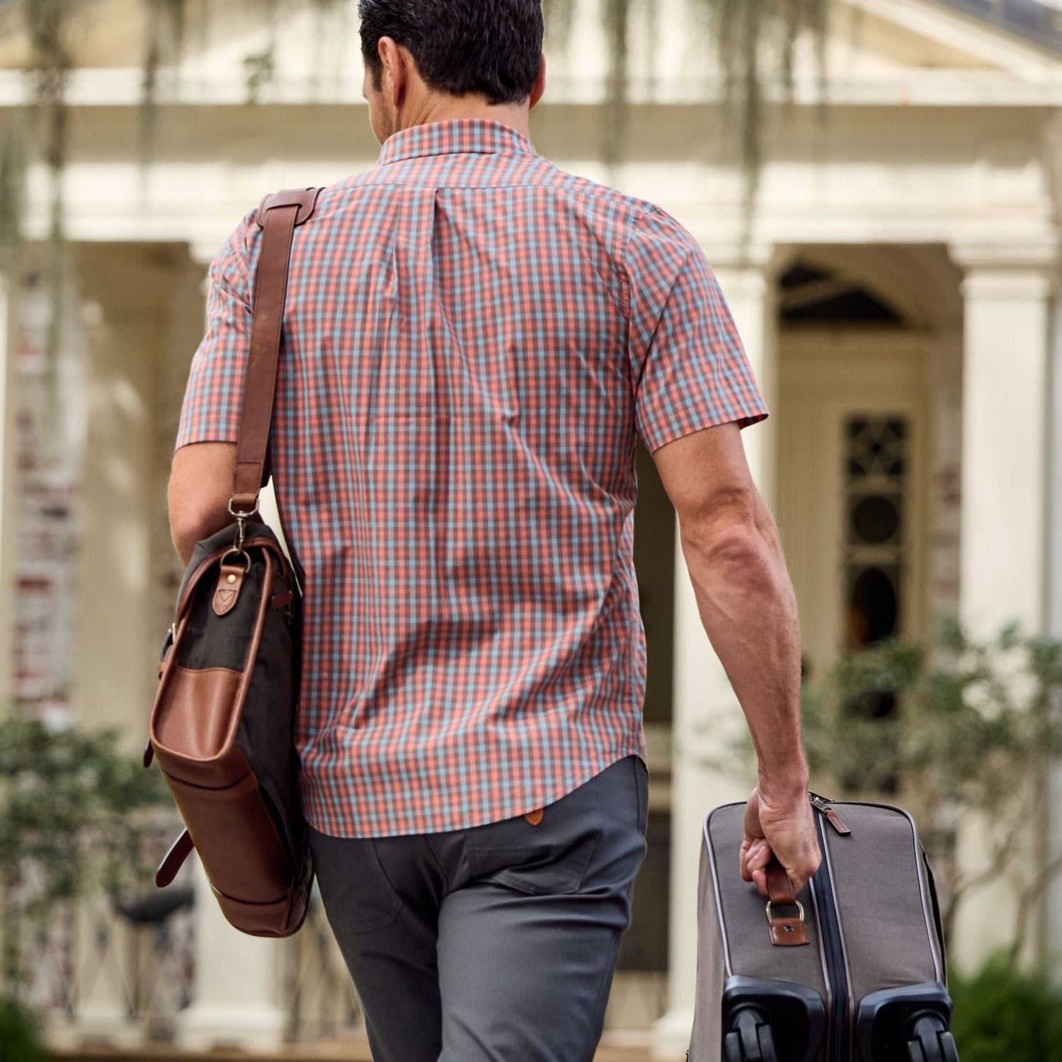 Man in a Tom Beckbe Tidewater Shirt, carrying a shoulder bag and rolling a suitcase on a path in front of a building.