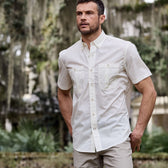 Man in a white Tidewater Shirt by Tom Beckbe and beige pants, standing outdoors with trees and greenery in the backdrop.