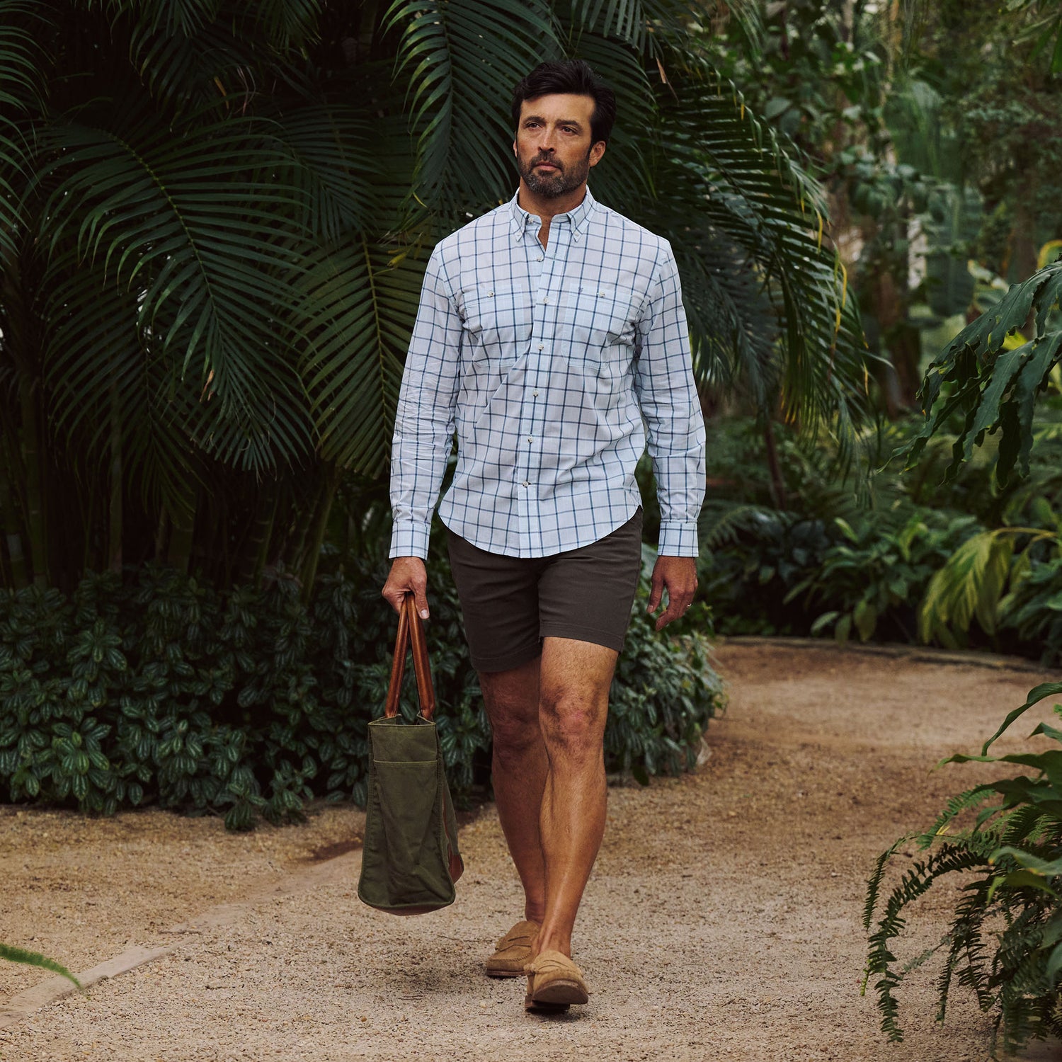 Man walking in a garden, wearing the Tom Beckbe Tidewater Shirt, shorts, loafers, and carrying a green bag.