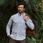 Man wears Tom Beckbe Tidewater Shirt, standing outdoors with a green tote amid lush greenery.