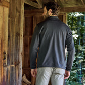 Man in a dark Tucker Fleece Quarter Zip by Tom Beckbe faces away from a rustic cabin, with trees in the background.