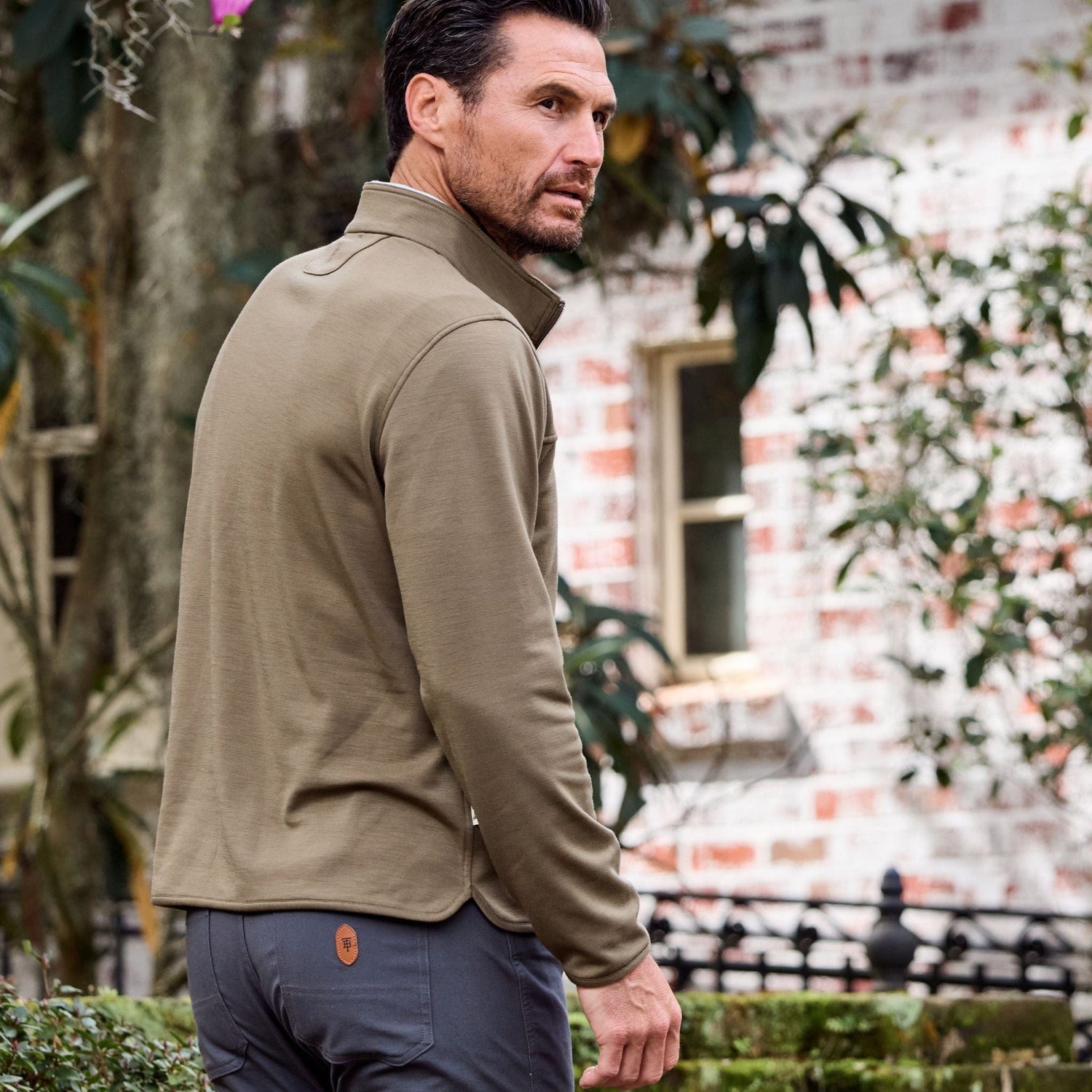 Man in an olive Tom Beckbe Tucker Fleece Quarter Zip and dark pants near foliage outside a brick building.