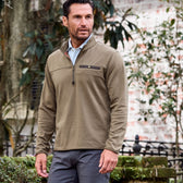 Man in a Tom Beckbe Tucker Fleece Quarter Zip stands outdoors amidst greenery and a brick wall.