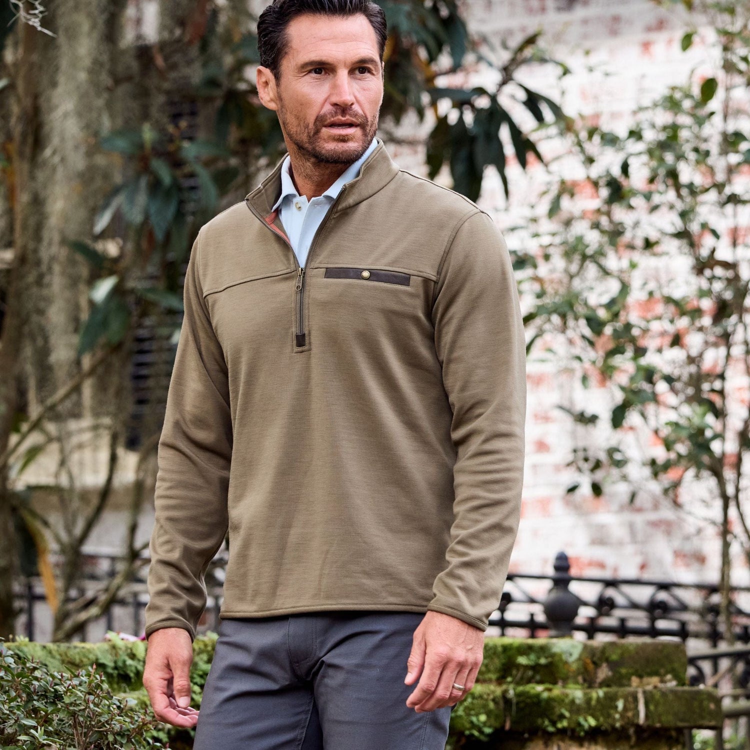 Man in a Tom Beckbe Tucker Fleece Quarter Zip stands outdoors amidst greenery and a brick wall.