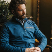 Man in Tom Beckbe Tucker Fleece Quarter Zip sits indoors with hands folded, gazing slightly to the side.