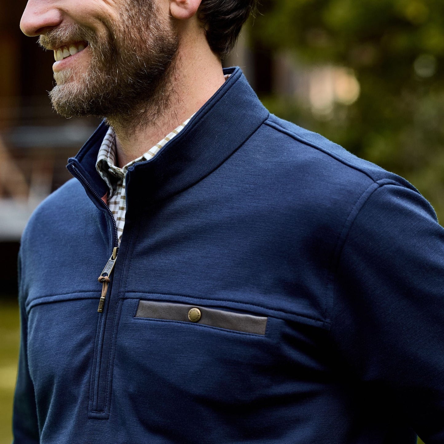 A man in a Tom Beckbe Tucker Fleece Quarter Zip, worn over a checkered shirt, stands outdoors and smiles warmly.