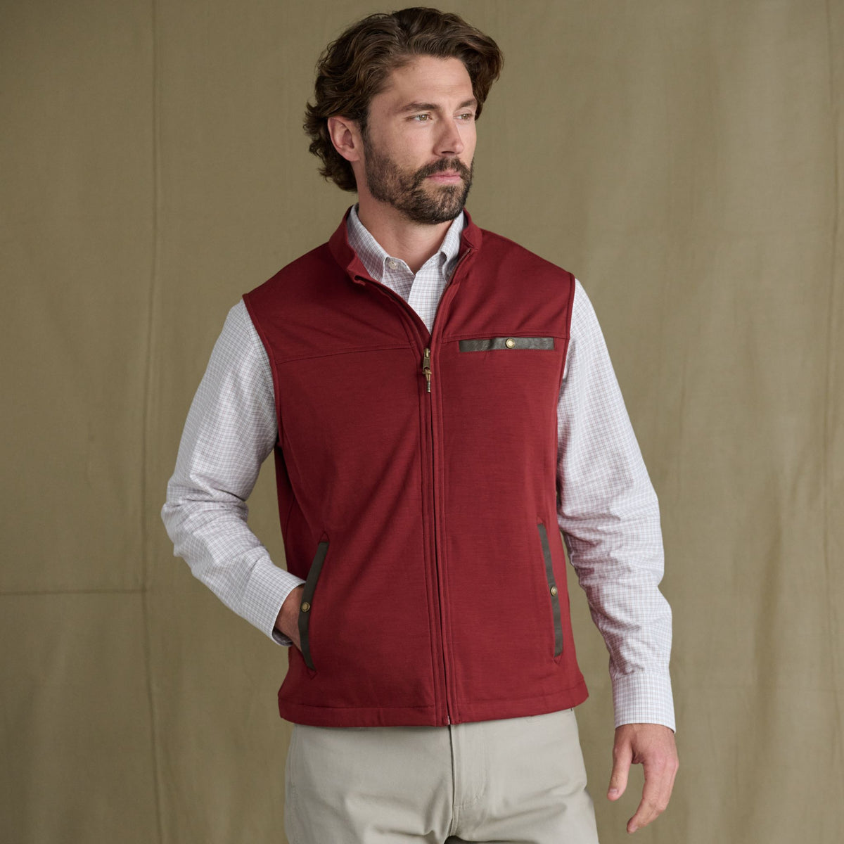 Man wears Tom Beckbe Tucker Fleece Vest over a checkered shirt and tan pants, standing before a plain brown backdrop.