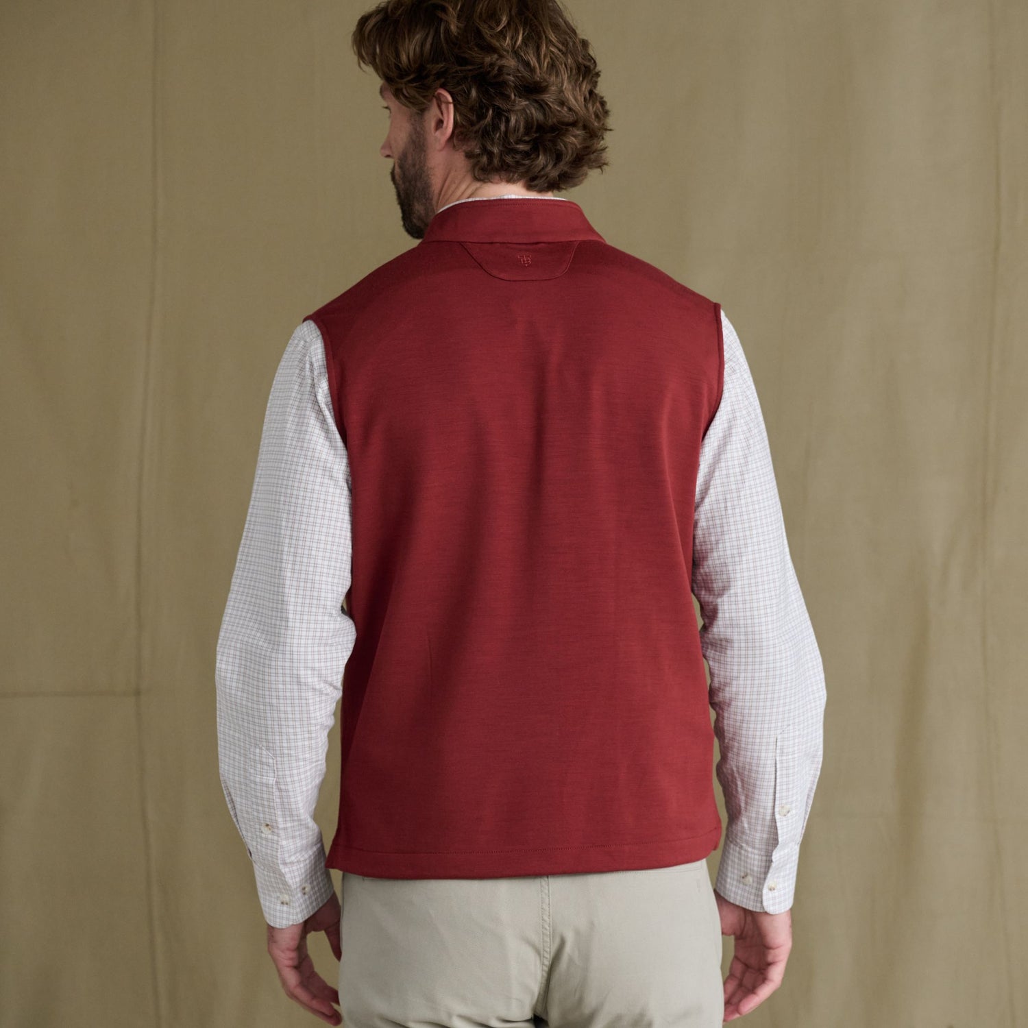 Man faces away, wearing a Tom Beckbe Tucker Fleece Vest over a checked shirt and beige pants.