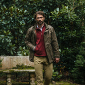 Man in a Tom Beckbe Tucker Fleece Vest stands in a garden with lush greenery and a stone bench behind him.