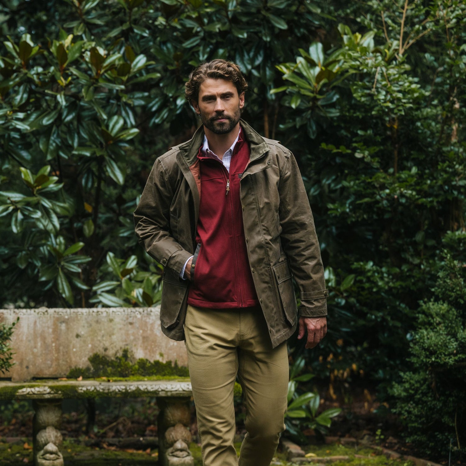 Man in a Tom Beckbe Tucker Fleece Vest stands in a garden with lush greenery and a stone bench behind him.