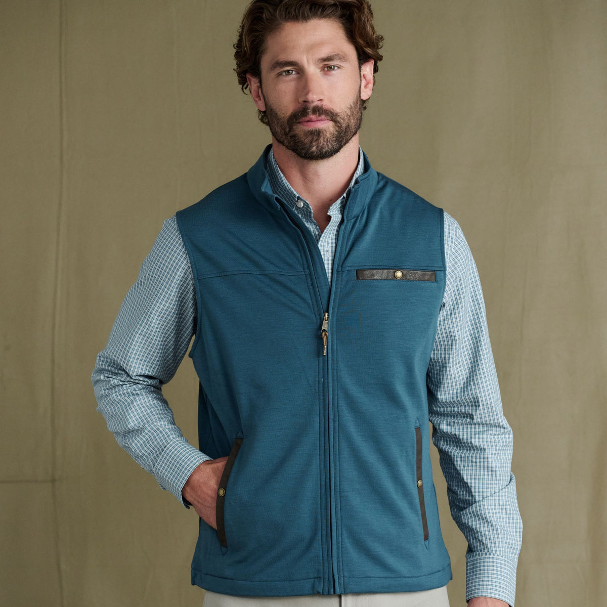 Man wearing a Tom Beckbe Tucker Fleece Vest over a checked shirt, hand in pocket, against a plain background.