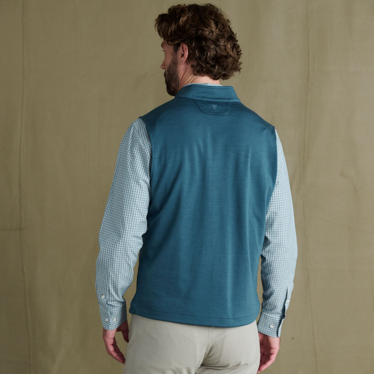Man wearing a Tom Beckbe Tucker Fleece Vest over a checkered shirt and beige pants, standing against a plain backdrop.
