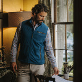 Man in a Tom Beckbe Tucker Fleece Vest and plaid shirt stands by a window, looking down at papers on a wooden desk.