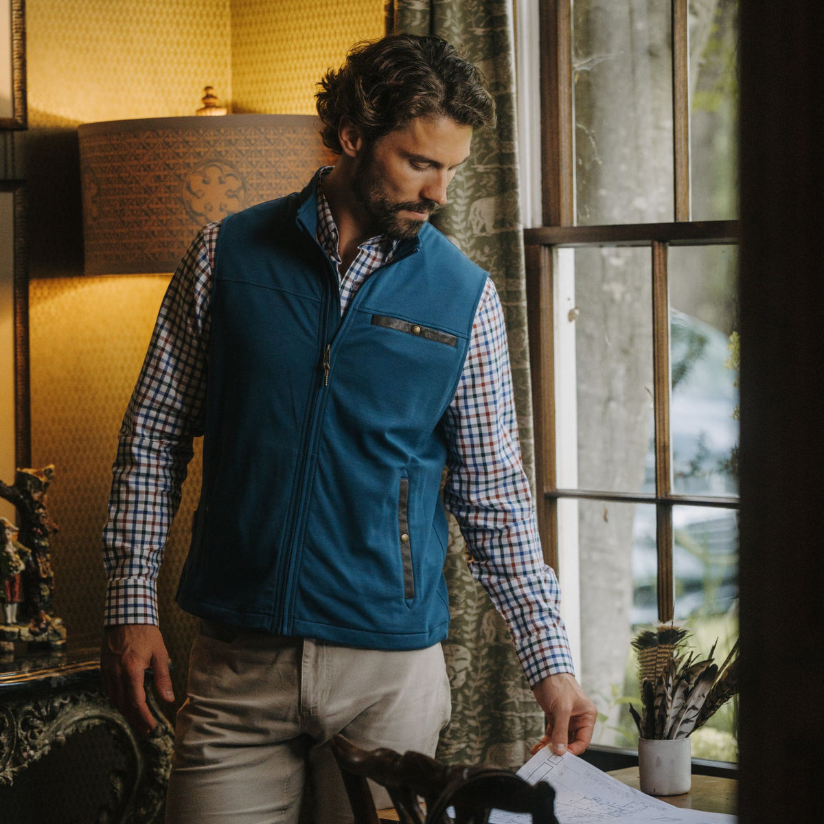 Man in a Tom Beckbe Tucker Fleece Vest and plaid shirt stands by a window, looking down at papers on a wooden desk.
