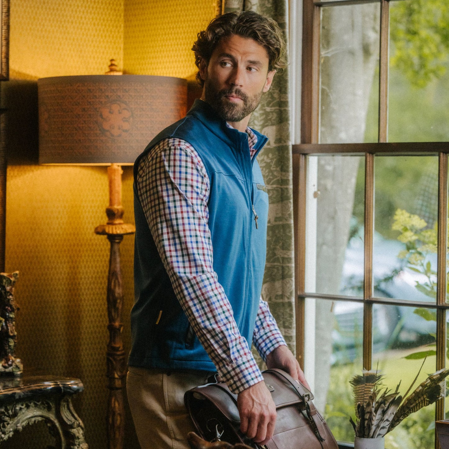 Man in a blue Tucker Fleece Vest by Tom Beckbe and plaid shirt stands by a window, holding a brown leather bag.